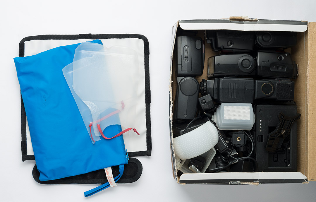 Main flash box with top layers of flash bouncer, milk bottle diffusers and pack of filter gels removed. In the box are speedlight flashes, various flash accessories, and a small LED light panel.